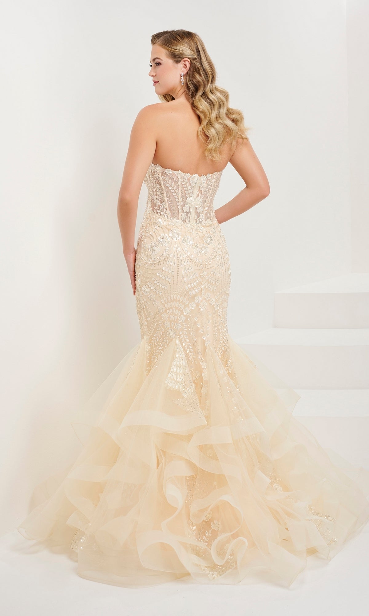 A woman stands with her back to the camera in the Tiffany Strapless Beaded Mermaid Prom Dress 16103, featuring beaded floral designs, lace and sequin details, and a ruffled tulle skirt.